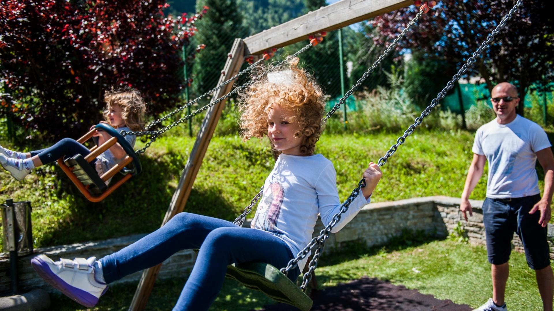 Bambini che giocano sull'altalena, sorvegliati da un adulto in un parco soleggiato.