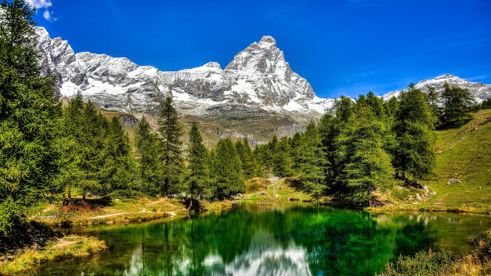 Lago alpino con riflessi montani, circondato da alberi e cime innevate.