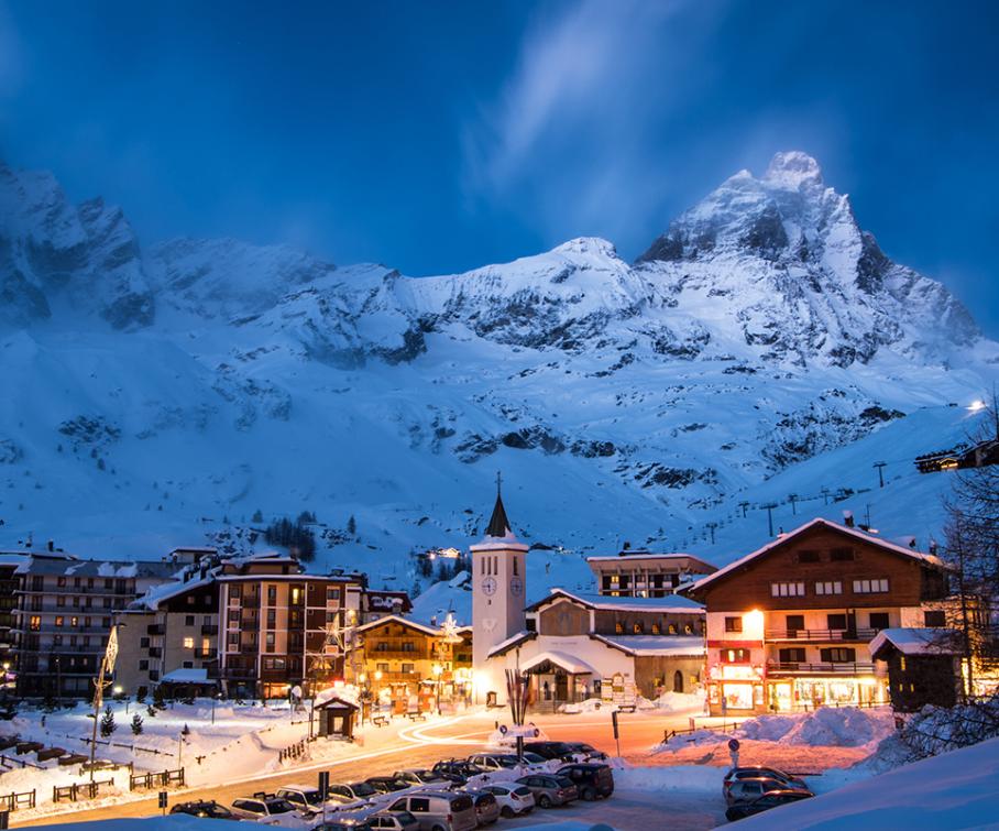 Villaggio innevato di montagna con case illuminate e montagne sullo sfondo al crepuscolo.